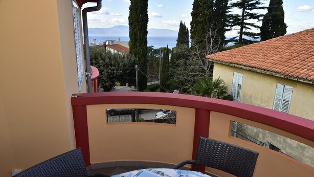 A balcony with a round table and two chairs, overlooking a coastal area with buildings and trees.