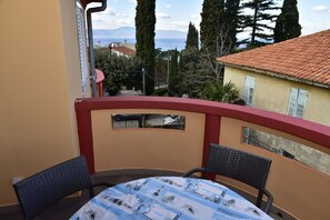 Un balcon avec une table ronde et deux chaises, donnant sur une zone côtière avec des bâtiments et des arbres.