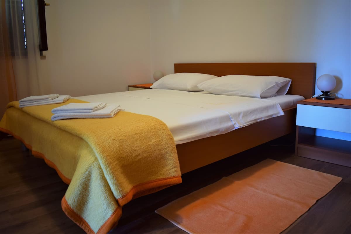 A neatly made bed with white linens and a yellow throw blanket, a bedside table with a lamp, and a wooden floor.