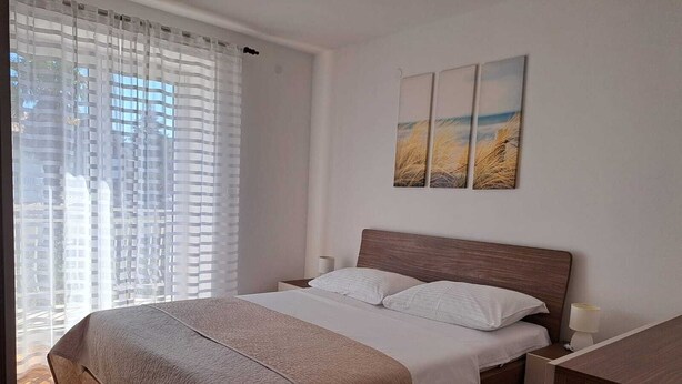 A bedroom with a bed, a nightstand, a desk, and a window with sheer curtains.