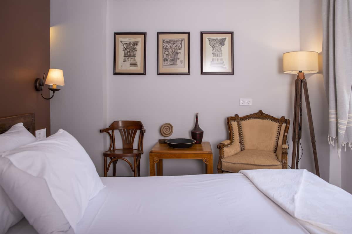 Ein Hotelzimmer mit einem Bett, zwei Stühlen, einem kleinen Tisch mit Schale und Vase sowie drei gerahmten Bildern an der Wand.