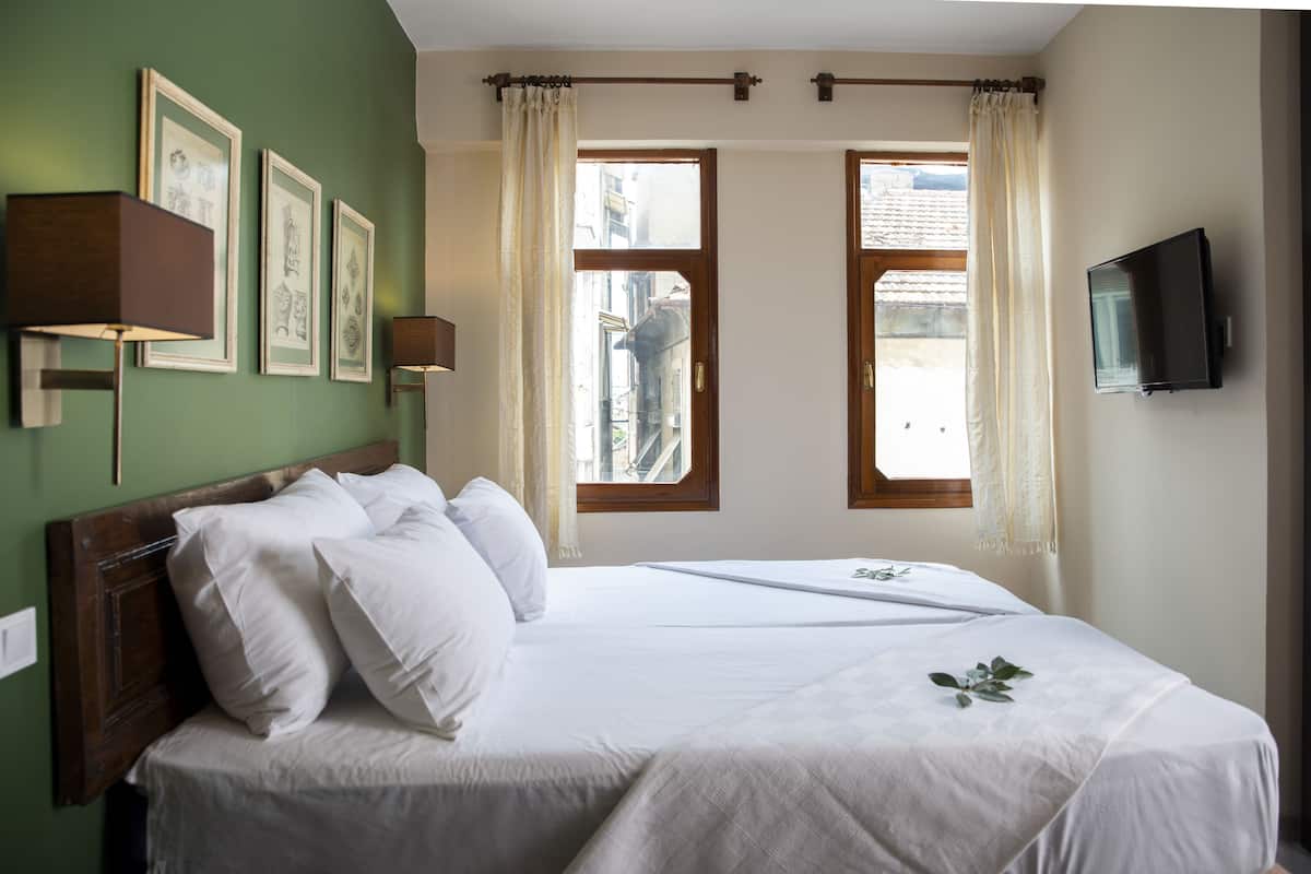 Ein Hotelzimmer mit einem großen Bett, zwei Fenstern mit Vorhängen, einem an der Wand montierten Flachbildfernseher und gerahmten Bildern.