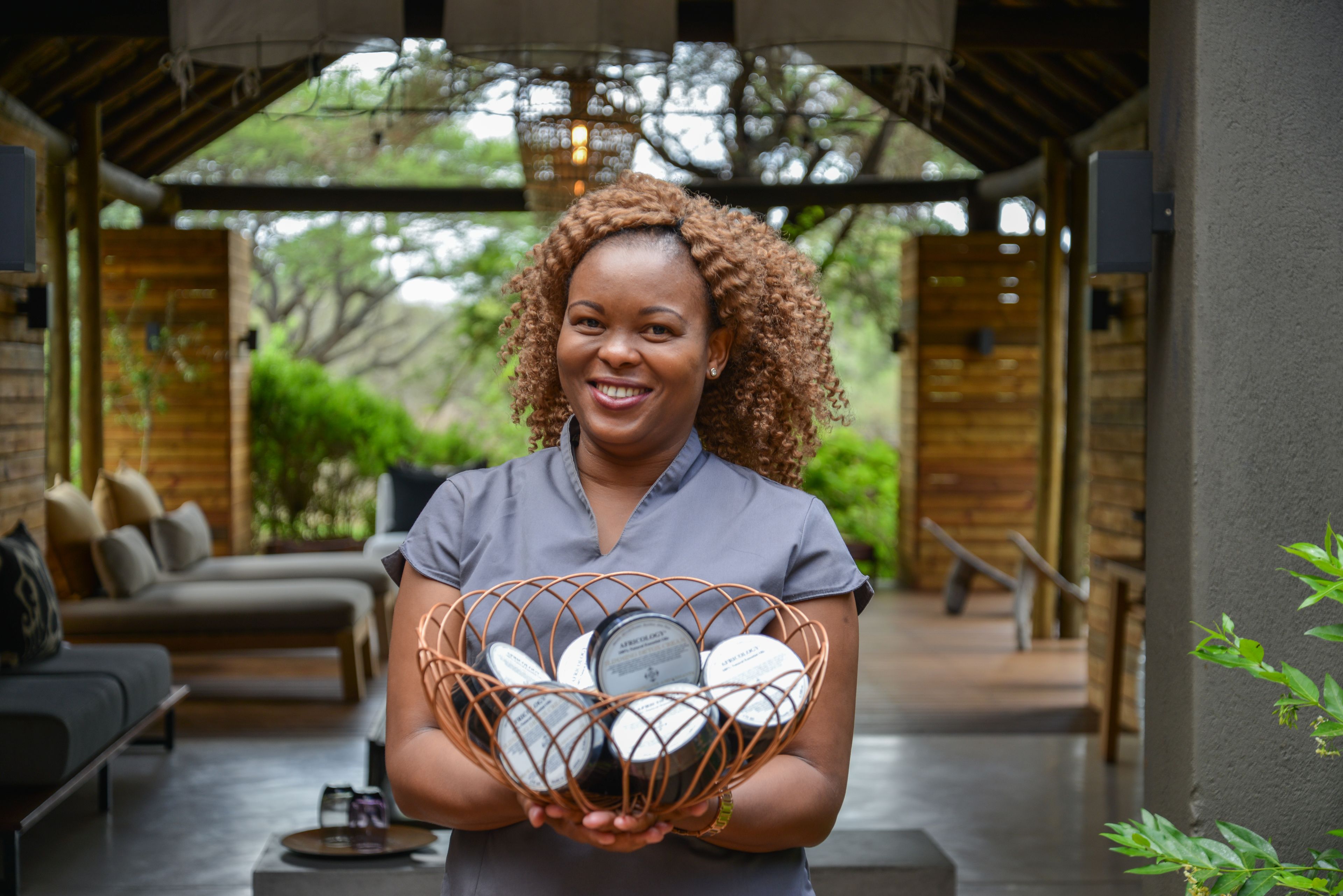 A person holding a basket of wine bottles in a covered outdoor area with wooden furniture and greenery in the background.