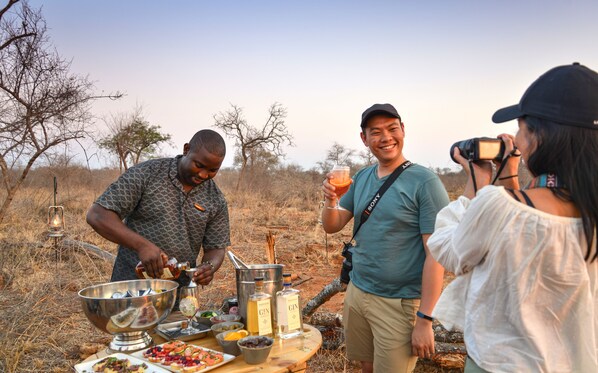 A group of people outdoors with a barbecue setup, including a metal bowl, bottles, and food.
