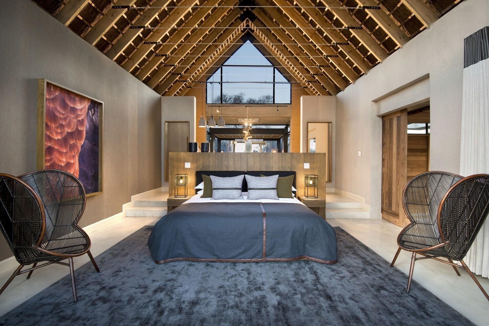 A bedroom with a large bed, two chairs, a wooden ceiling, and a view through a window.