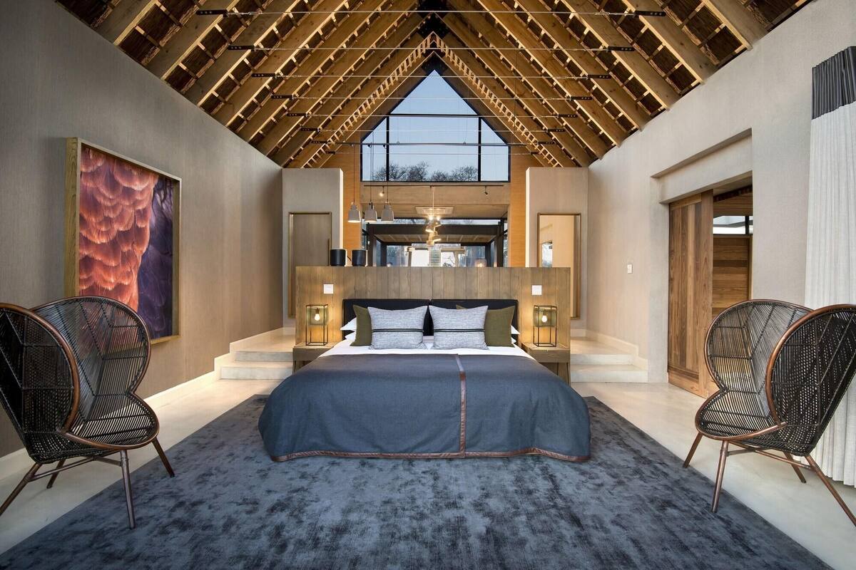 Ein Schlafzimmer mit einem großen Bett, zwei Stühlen, einer Holzbalkendecke und Blick durch ein Fenster.