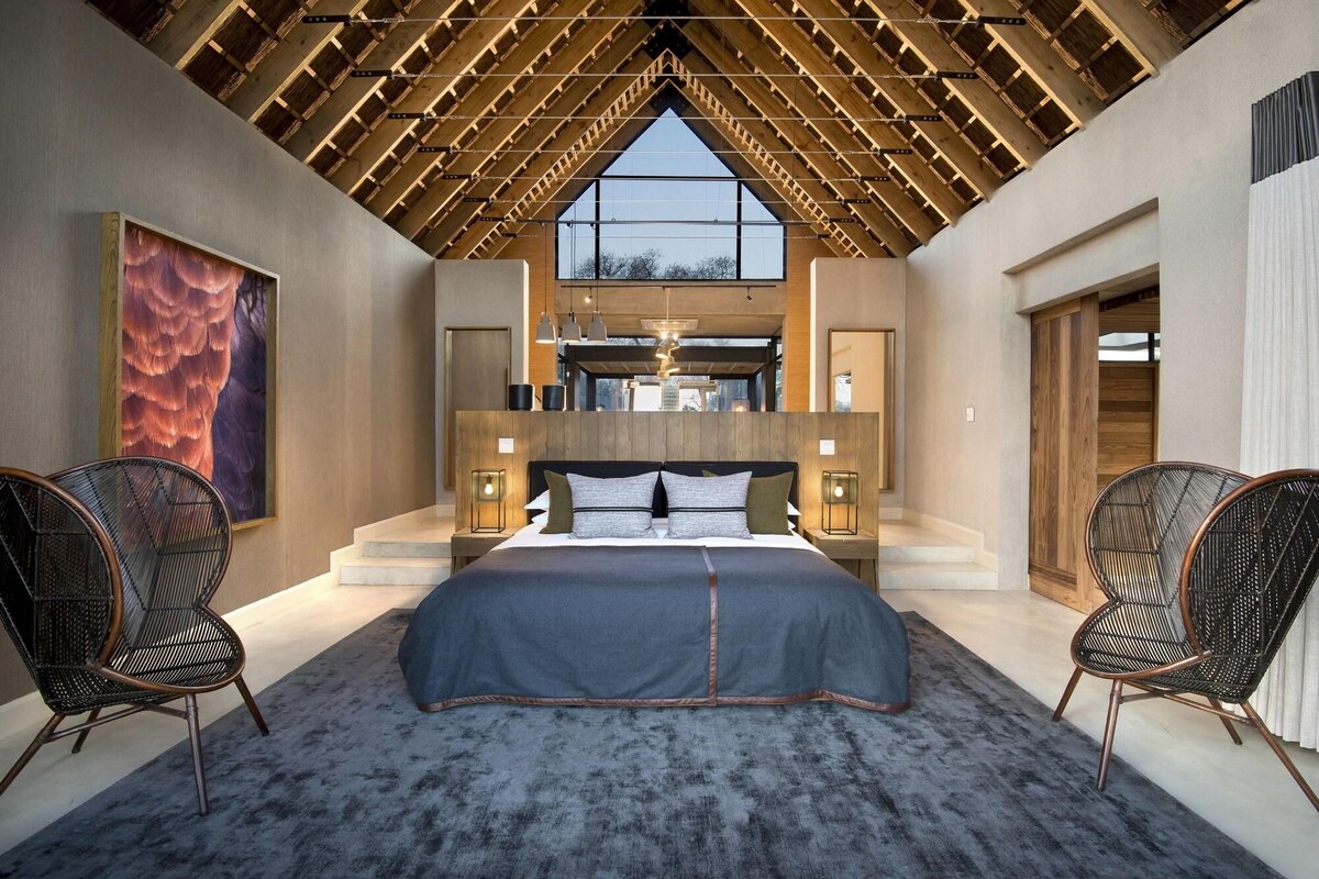A bedroom with a large bed, two chairs, a wooden ceiling, and a view through a window.