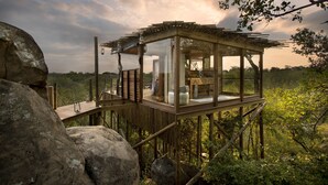 A wooden viewing platform with glass walls, surrounded by rocks and vegetation, overlooking a natural landscape.