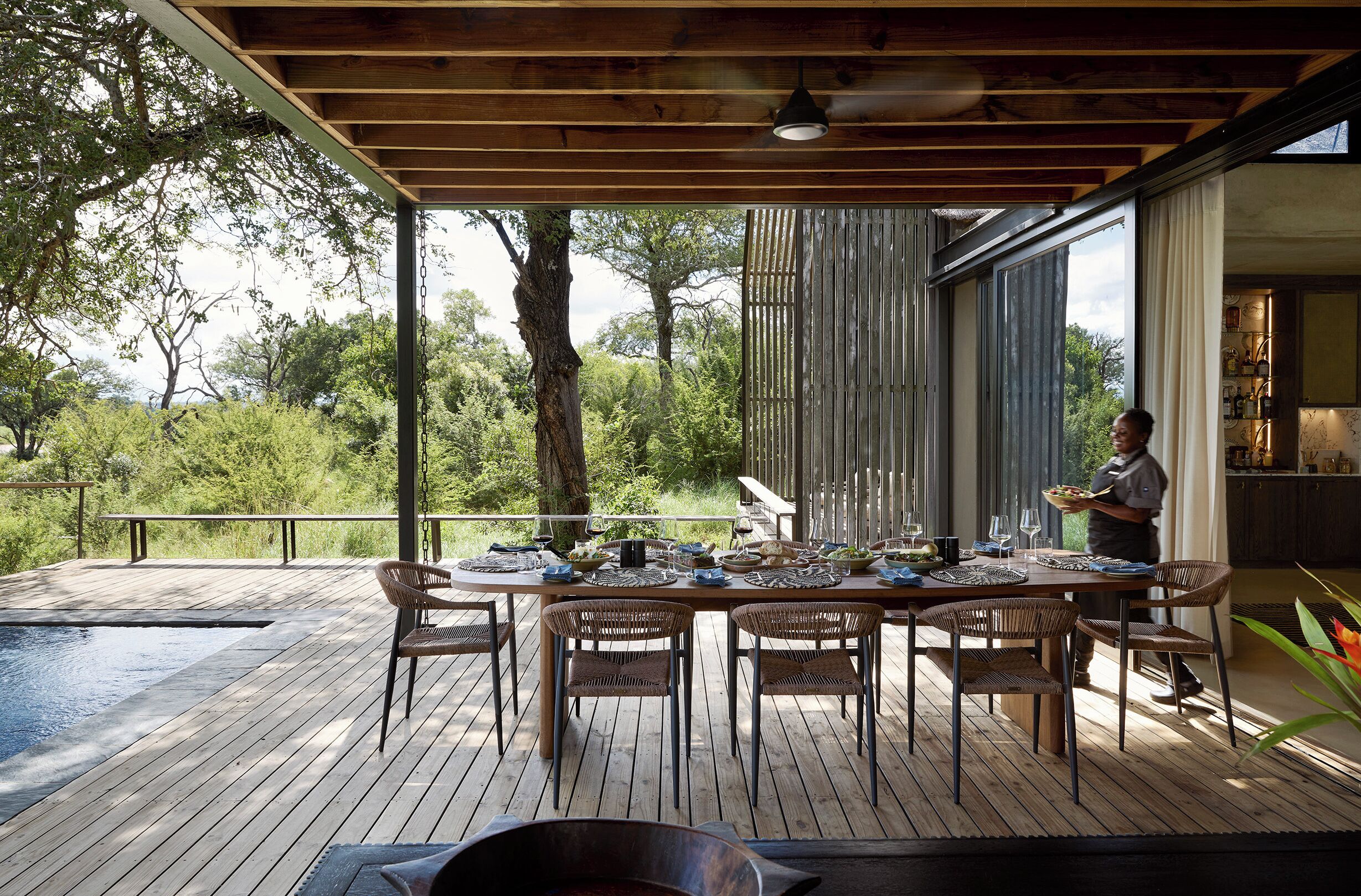 Een houten terras met een eettafel voor zes personen, omringd door rieten stoelen, met uitzicht op een zwembad en weelderige begroeiing.