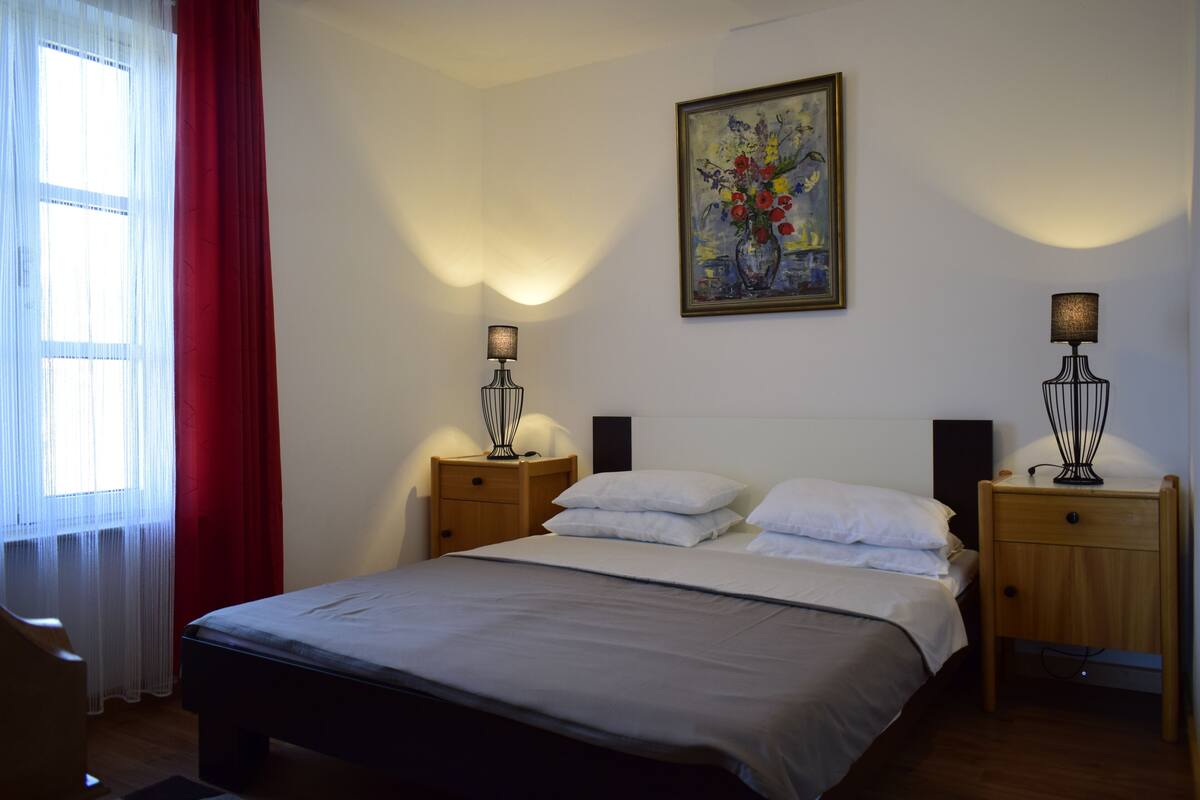 A neatly made bed with white linens, two bedside tables with lamps, a painting on the wall, and a window with red curtains.