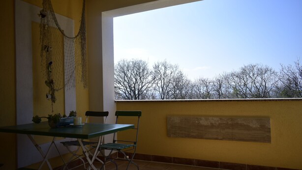 A balcony with a table and chairs, a view of trees, and a yellow wall. - Apartments Mermaid (Malinska-Dubasnica)