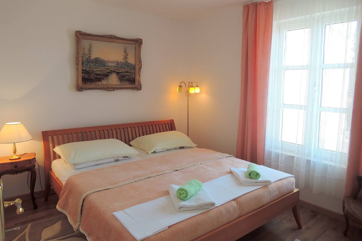 A neatly made bed with pillows, a bedside lamp, a painting on the wall, and a window with curtains.