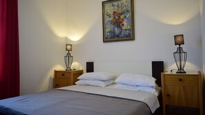 A neatly made bed with white linens, two bedside tables with lamps, a painting on the wall, and a red curtain on the left. - Apartments Mermaid (Malinska-Dubasnica)