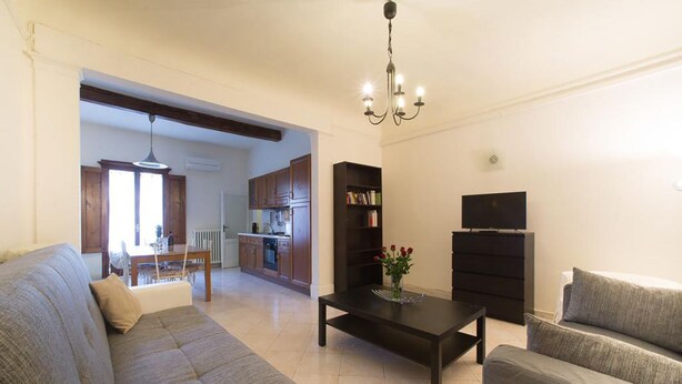 Una sala de estar con sofá, mesa de centro y mueble para la televisión. Se observa una cocina al fondo, con gabinetes de madera y un comedor.
