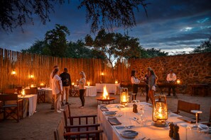 Un área de comedor al aire libre con mesas dispuestas para comensales, rodeada por un rústico muro de piedra y una valla de madera.