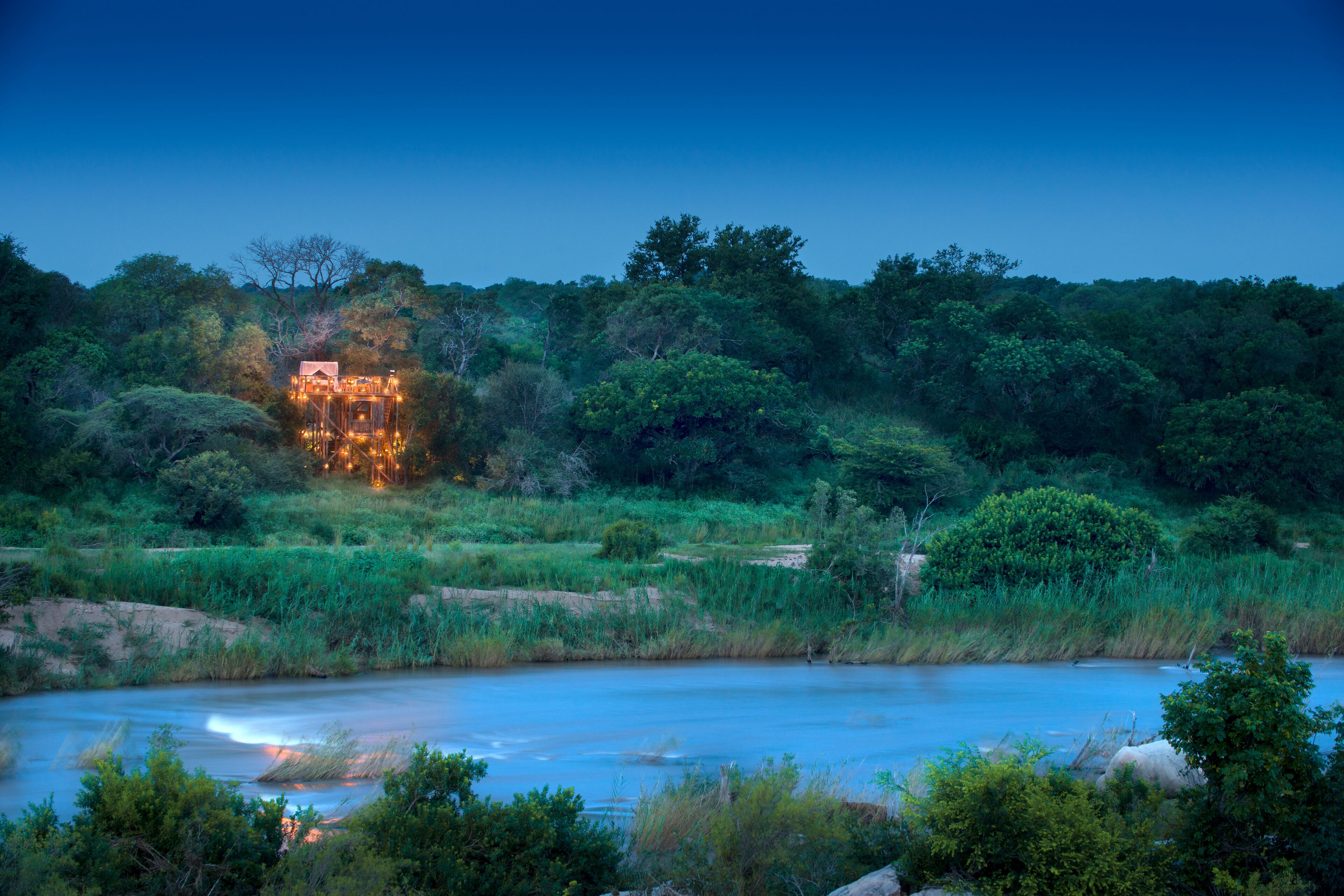 A riverside view with a building illuminated by warm lights, surrounded by dense greenery and trees.