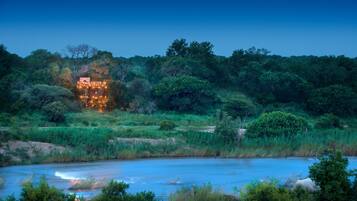 A riverside view with a building illuminated by warm lights, surrounded by dense greenery and trees.