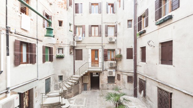 Ein schmaler Innenhof mit einer zentralen Treppe, die zu einer oberen Etage führt. An den Seiten befinden sich Gebäude mit zahlreichen Fenstern und Fensterläden, einige mit Pflanzen.