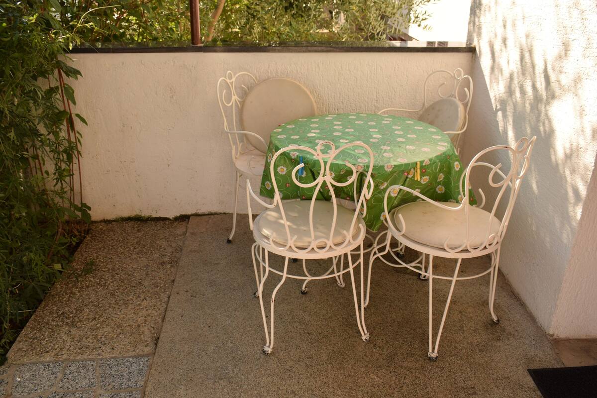 A small outdoor patio with a round table and four white metal chairs, surrounded by a low white wall and greenery.