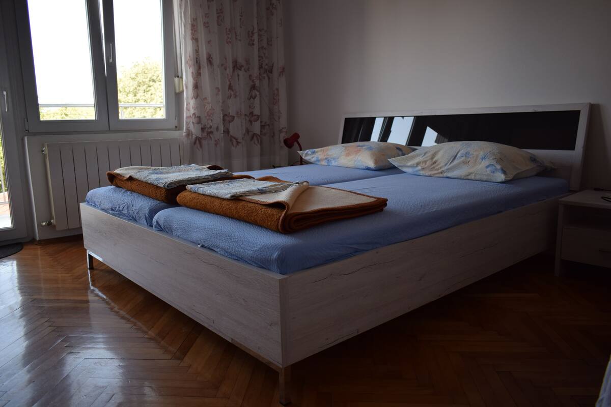 A bedroom with a wooden floor, a bed with a blue mattress and white headboard, and a window with floral curtains.