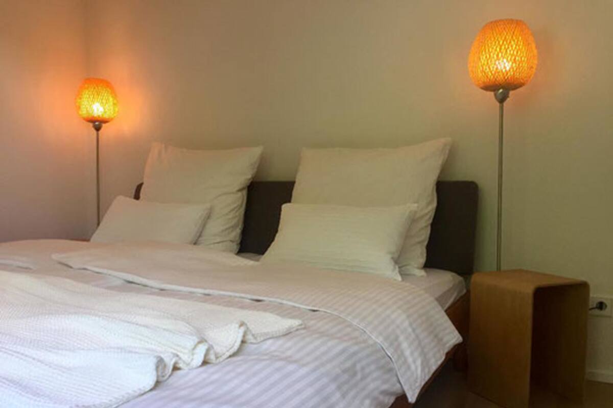 A neatly made bed with white linens and pillows, two floor lamps with orange shades, and a wooden nightstand.