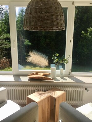 Una habitación acogedora con un ventanal grande, una lámpara de fibra natural, una pequeña mesa de madera y un radiador abajo.