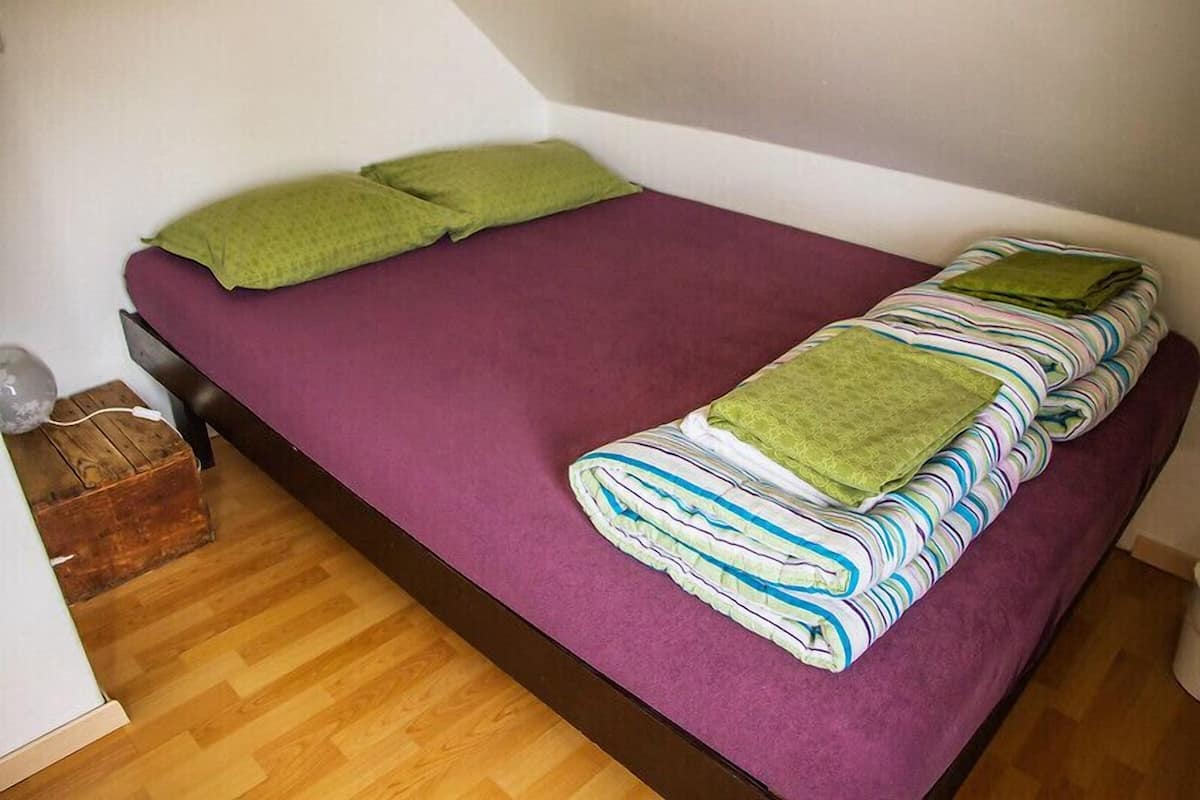 Een kleine eenpersoonskamer met een paars dekbed, twee groene kussens en een netjes opgevouwen gestreept deken.