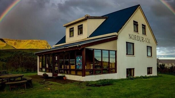 A building named 'Nordur-Vik' with a large glass facade, a covered outdoor seating area, and a view of a rainbow in the background. - Vik HI hostel (Vik I Myrdal)