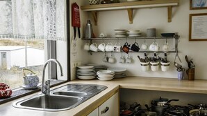 A kitchen with a stainless steel sink, a window with lace curtains, and a shelf with white mugs and plates. - Vik HI hostel (Vik I Myrdal)