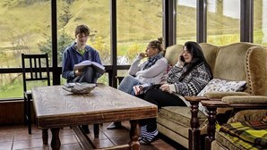 A cozy room with a large window offering a view of a green landscape. There is a wooden table and a sofa with cushions. - Vik HI hostel (Vik I Myrdal)