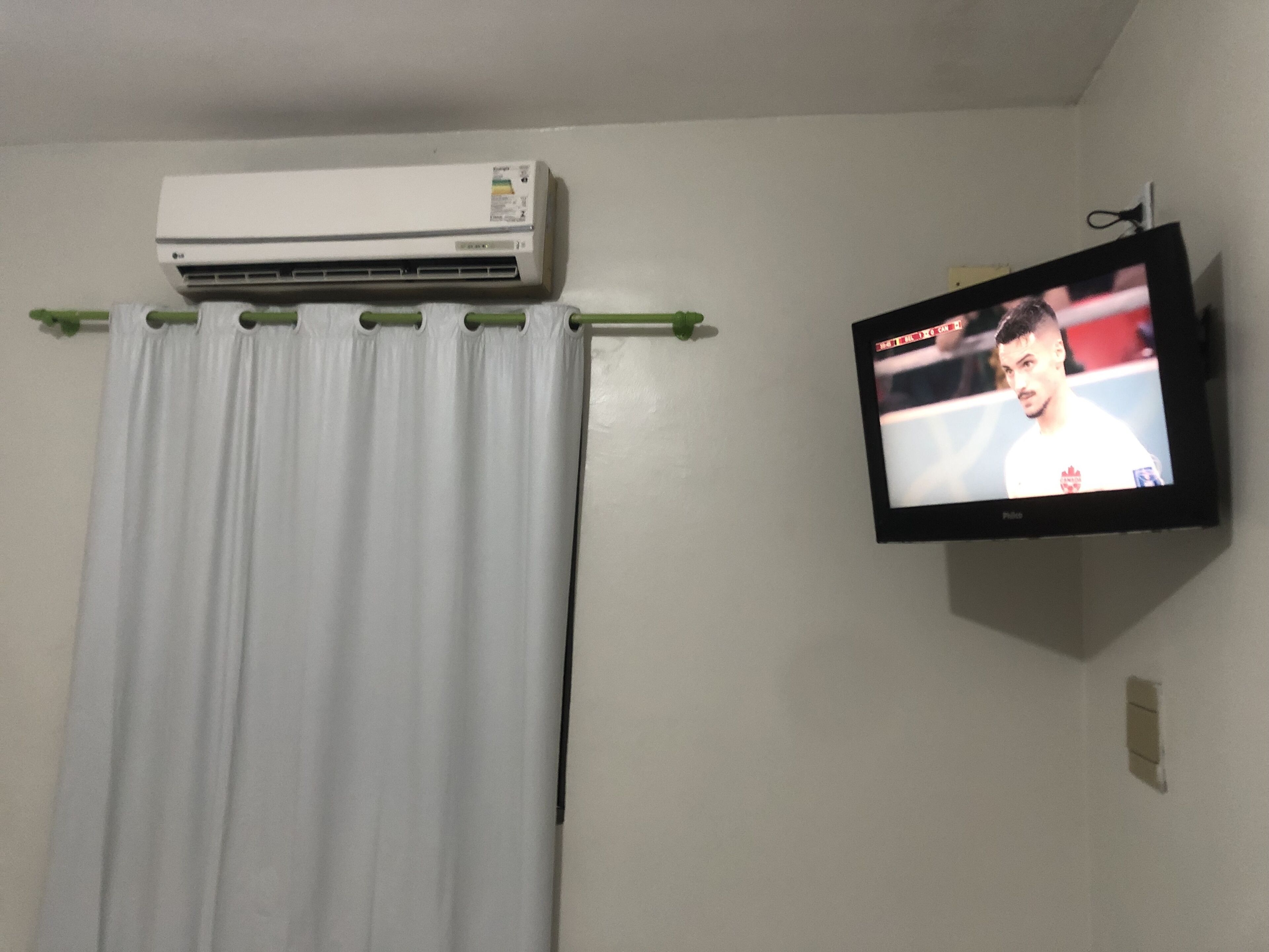 Una habitación con aire acondicionado blanco, cortina y televisor con un evento deportivo.