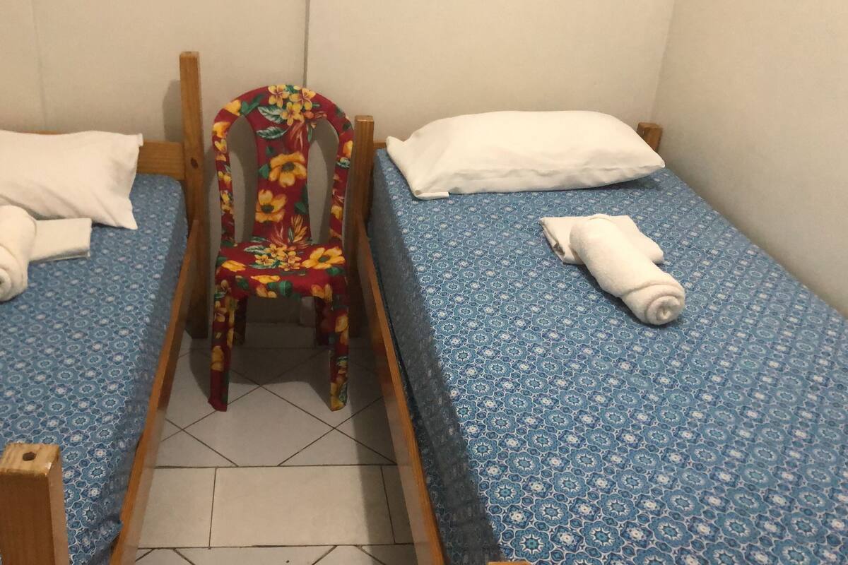 Ett litet rum med en enkel säng, en trästol i mitten, ett blått mönstrat överkast och vita kuddar.