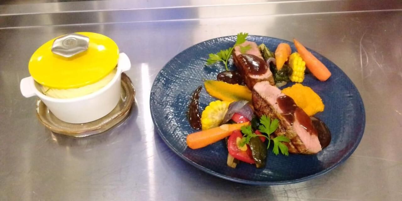 Un plato azul con un bistec, verduras y una olla con tapa amarilla sobre una superficie de acero inoxidable.