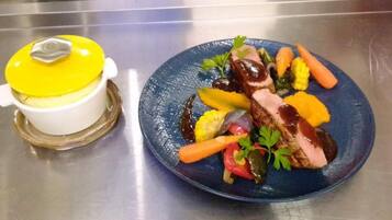 Un plato azul con un bistec, verduras y una olla con tapa amarilla sobre una superficie de acero inoxidable.