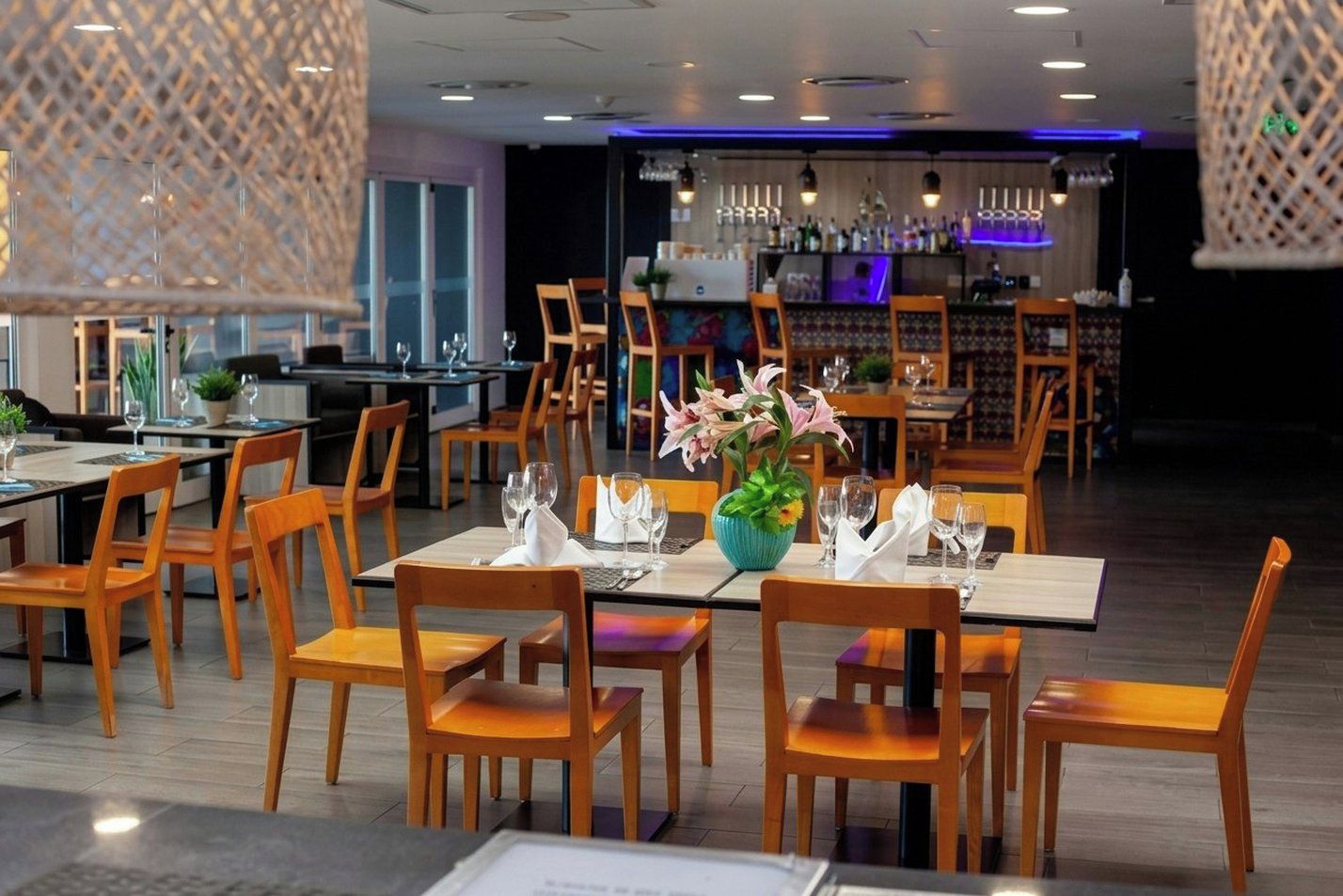 Ein Restaurant mit Holztischen und -stühlen, ein Barbereich im Hintergrund und hängenden Lampen.