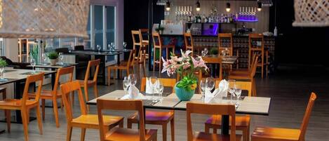 Ein Restaurant mit Holztischen und -stühlen, ein Barbereich im Hintergrund und hängenden Lampen.
