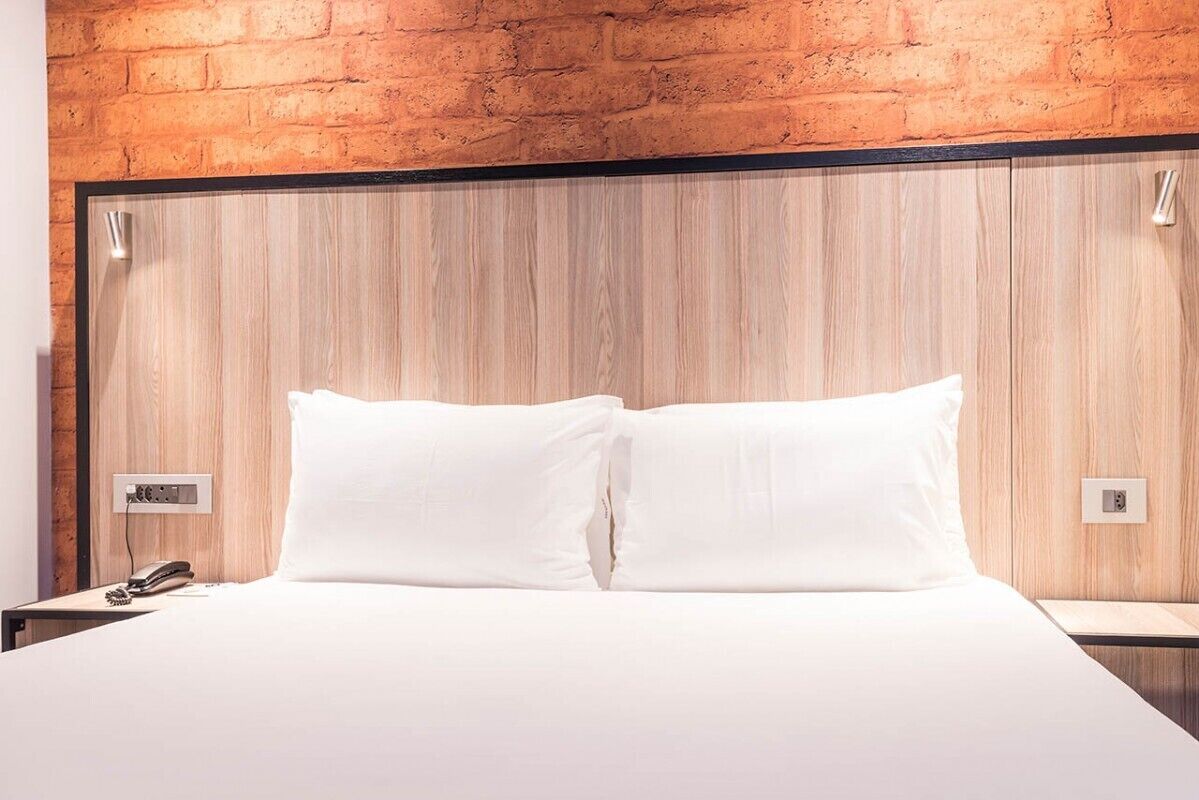 A hotel room with a wooden headboard, a bed with white linens, a bedside table with a phone and a lamp, and a brick wall.