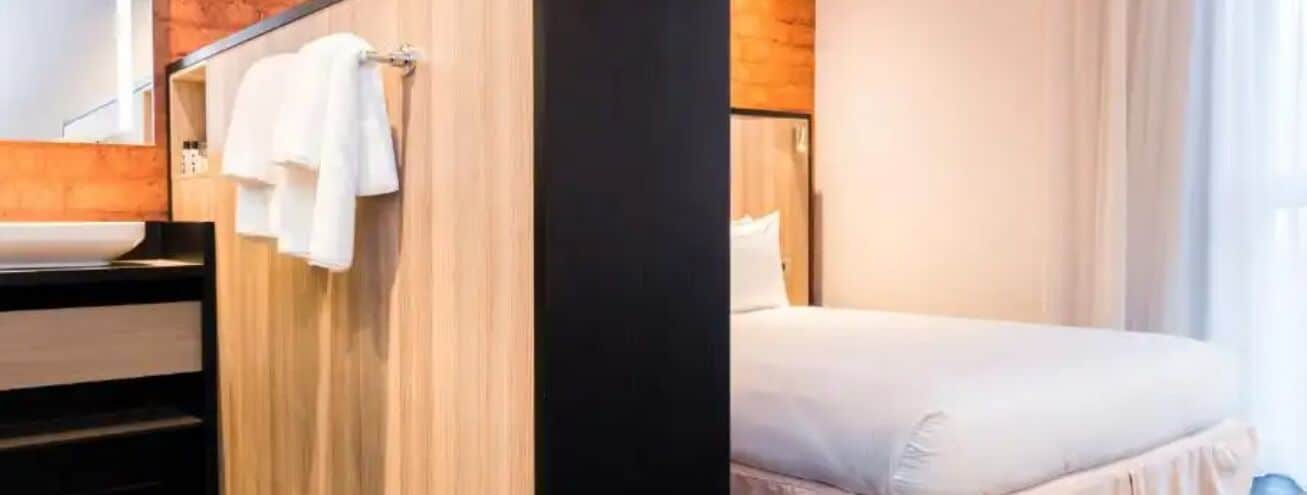 A hotel room with a bed, a wooden wall, and a bathroom with a sink and towel rack.