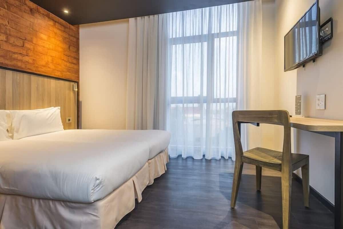 Quarto com uma cama grande, uma escrivaninha de madeira, uma cadeira, uma TV e uma janela com cortinas.