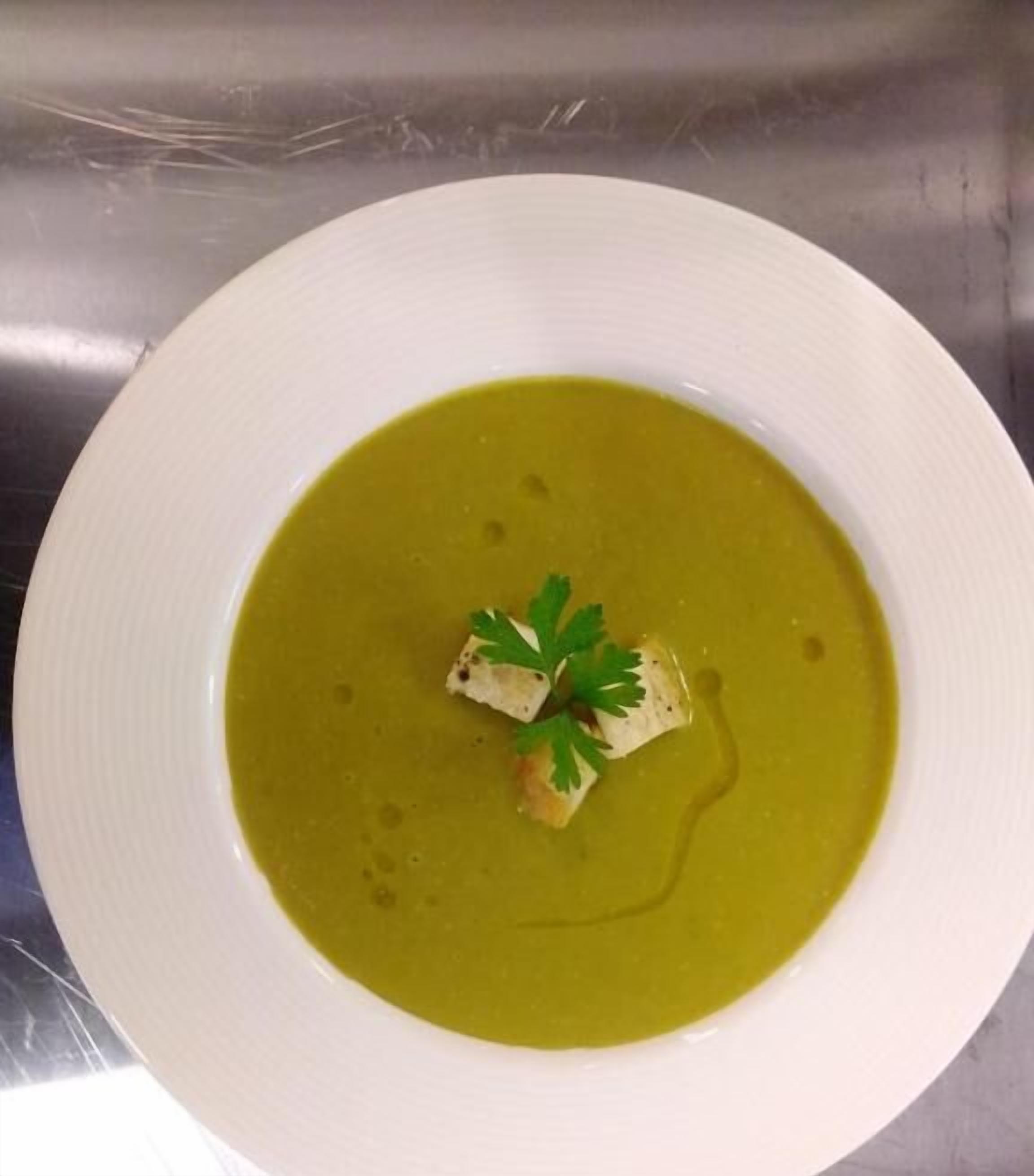Un tazón blanco con sopa verde, un trozo de pescado y perejil como guarnición, sobre una superficie de acero inoxidable.