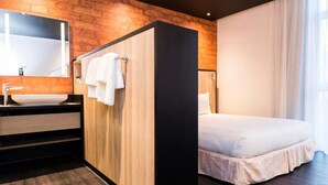 A modern hotel room with a wooden accent wall, a large bed, a bathroom with a sink and mirror, and a window with curtains. - ONOMO Hotel Maputo (Maputo)