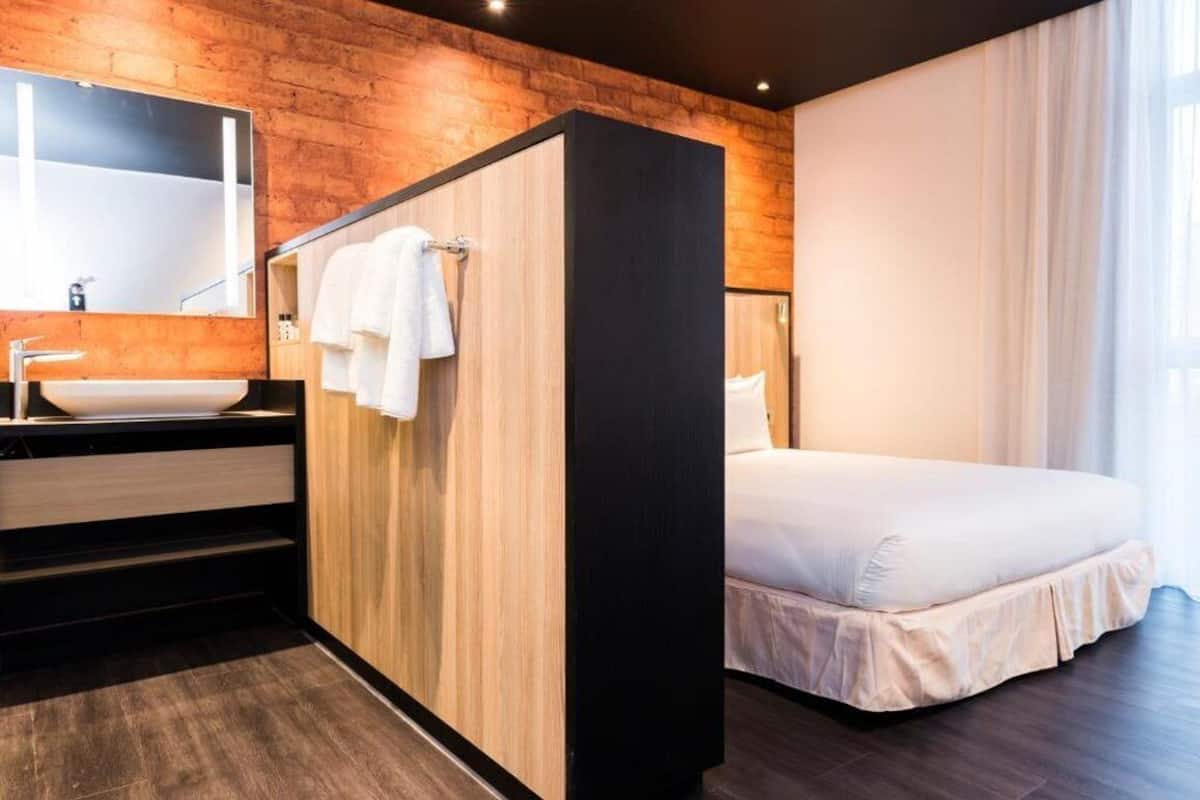 Quarto de hotel moderno com parede de destaque em madeira, uma cama grande, banheiro com pia e espelho, e janela com cortinas.