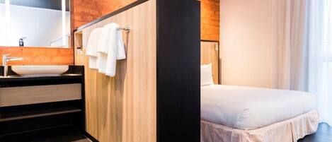 Una habitación de hotel moderna con un panel de madera decorativo, una cama grande, un baño con lavamanos y espejo, y una ventana con cortinas.