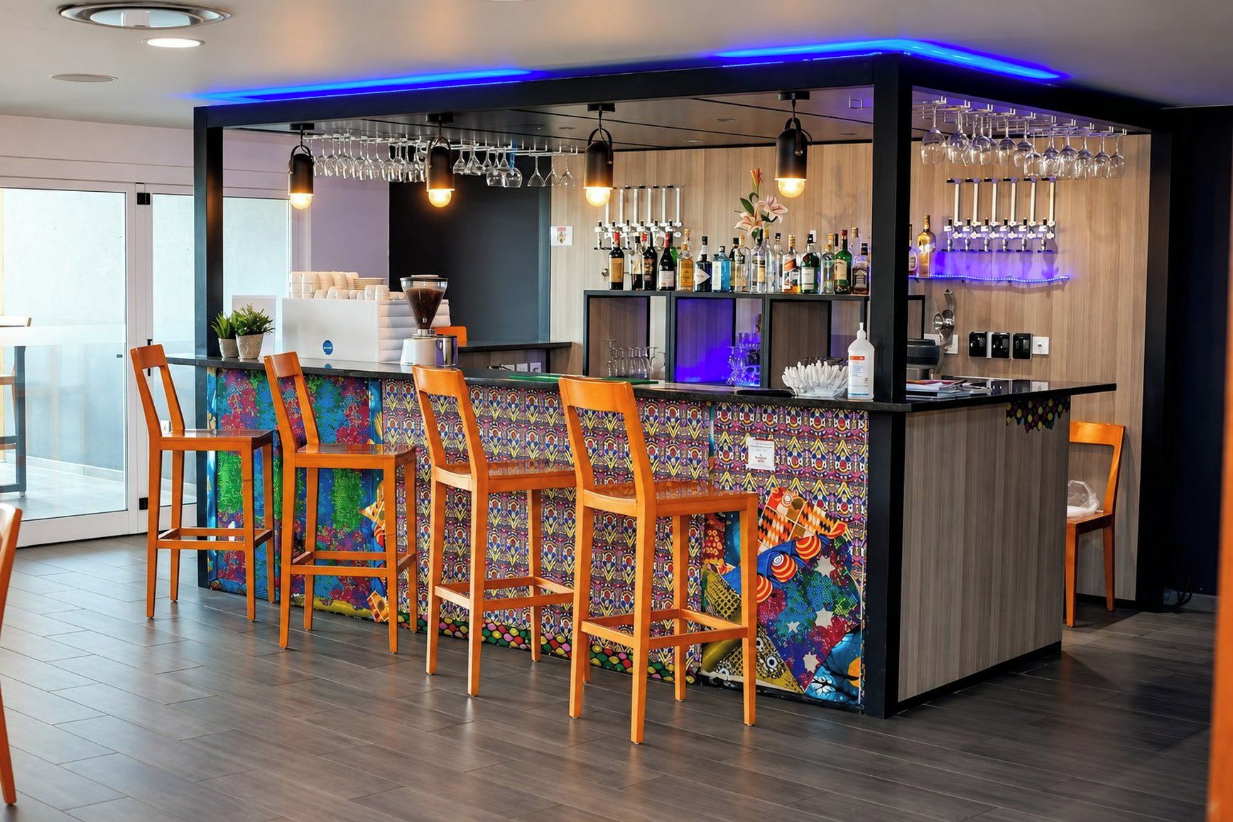 A bar area with a colorful patterned wall, wooden bar stools, and a variety of bottles and glasses.