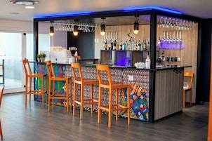 Una zona de bar con una pared de diseño colorido, taburetes de madera y una variedad de botellas y vasos.