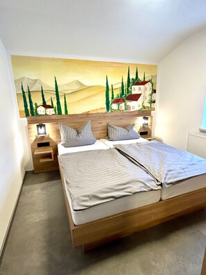 Una cama doble con estructura de madera, dos mesitas de noche con lámparas y un mural en la pared que representa un paisaje con árboles y edificios.