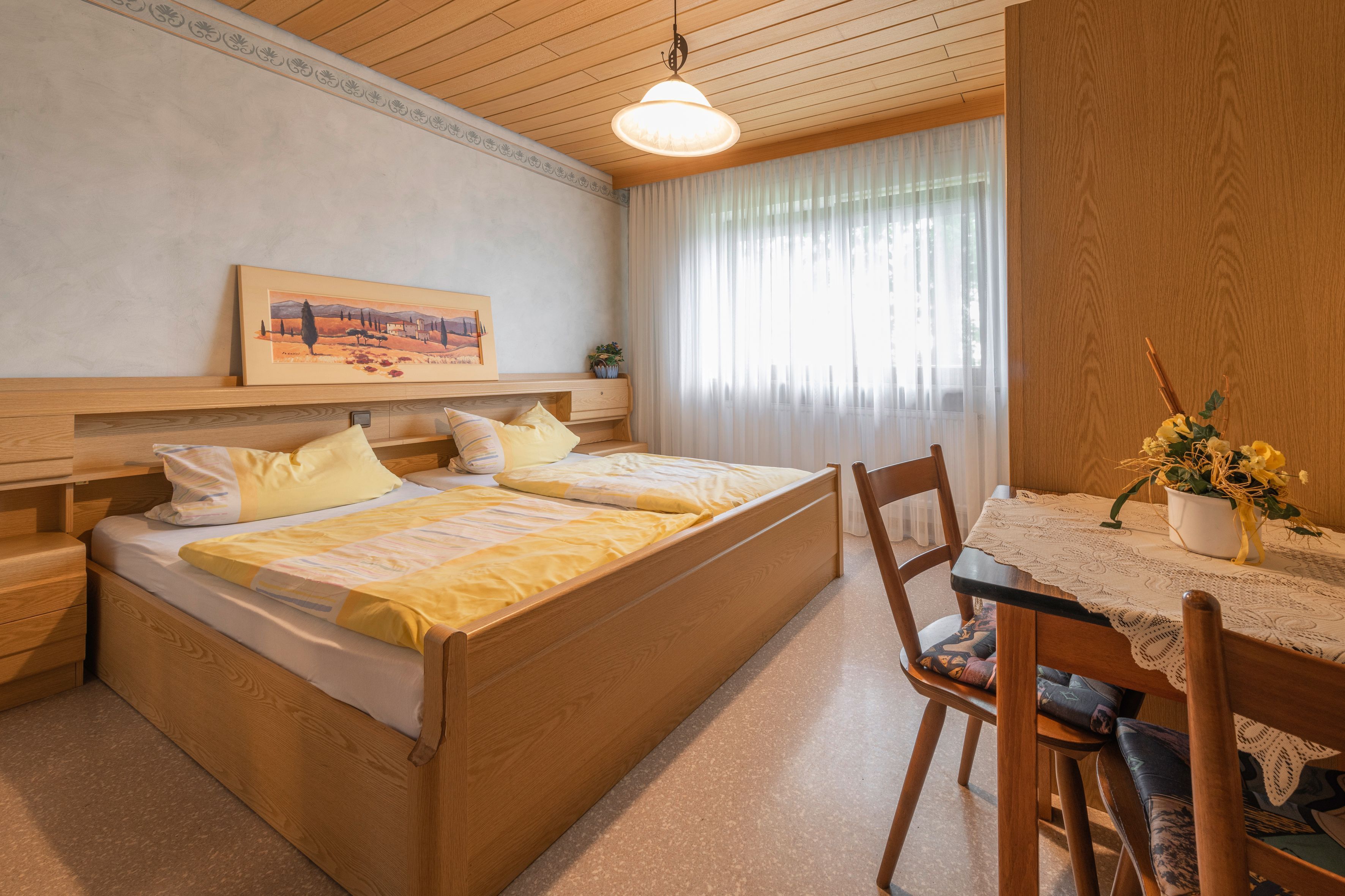 Una camera da letto con un letto di legno, comodini, un tavolino, una finestra con tende, un tavolo da pranzo con tovaglia in pizzo e un vaso con fiori.