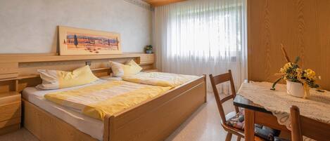 Una camera da letto con un letto di legno, comodini, un tavolino, una finestra con tende, un tavolo da pranzo con tovaglia in pizzo e un vaso con fiori.