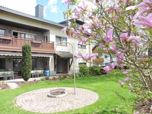 Un patio con un hogar circular de ladrillo, un alto pino y flores rosadas en flor.