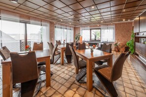 Un'area pranzo con tavoli e sedie in legno, ampie finestre e pavimento piastrellato.